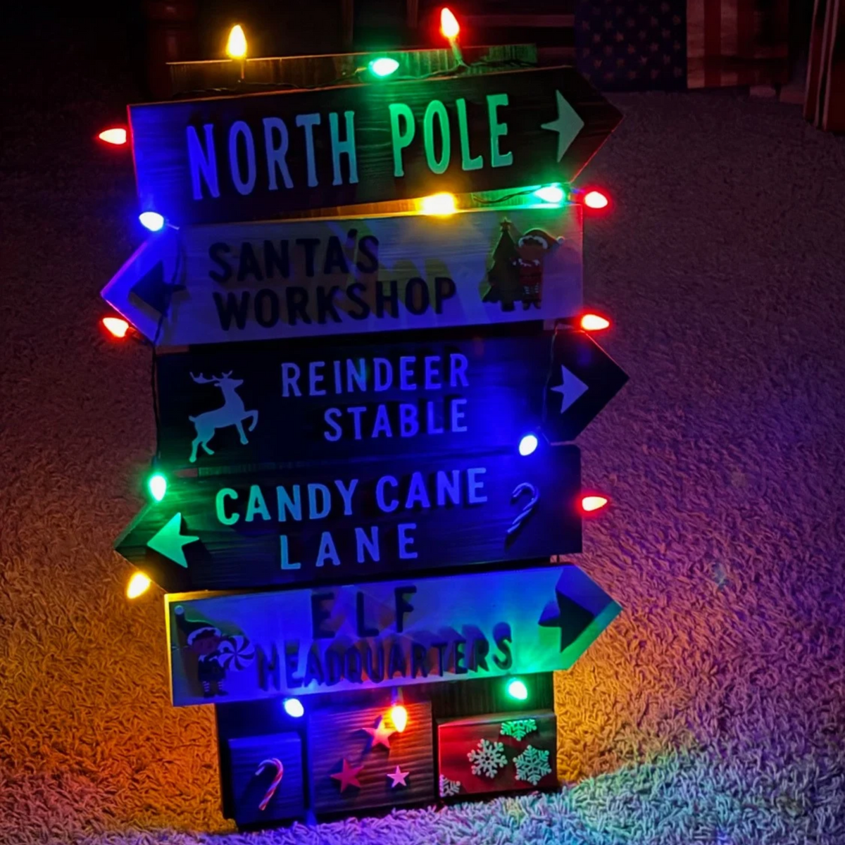 A rustic wooden Christmas signpost with five directional signs pointing North Pole, Santa's Workshop, Reindeer Stable, Candy Cane Lane, and Elf Headquarters, lit with multicolored string lights on a grassy surface.