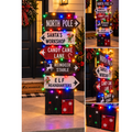 A festive lighted Christmas directional signpost decoration with multiple white arrow signs reading North Pole, Santa's Workshop, Candy Cane Lane, Reindeer stable, and Elf Headquarters,.  Lit with multicolored holiday string lights and decorated with small reindeer and elf graphics. 