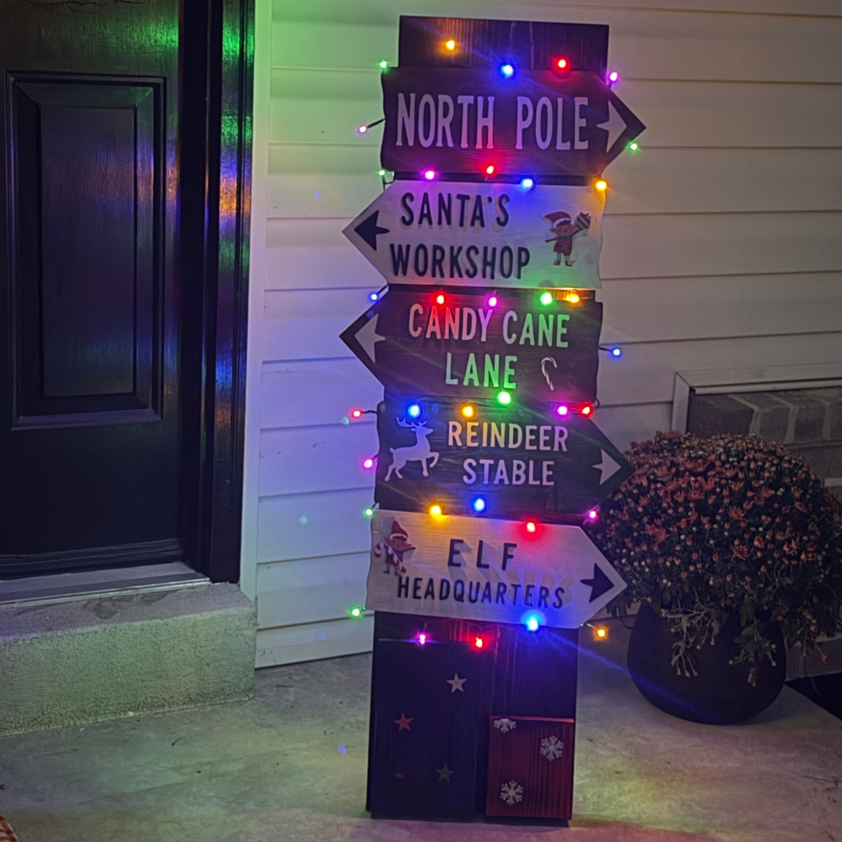 A tall decorative lighted wooden signpost with five Christmas-themed directional signs reading North Pole, Santa's Workshop, Candy Cane Lane, Reindeer Stable, and Elf Headquarters.