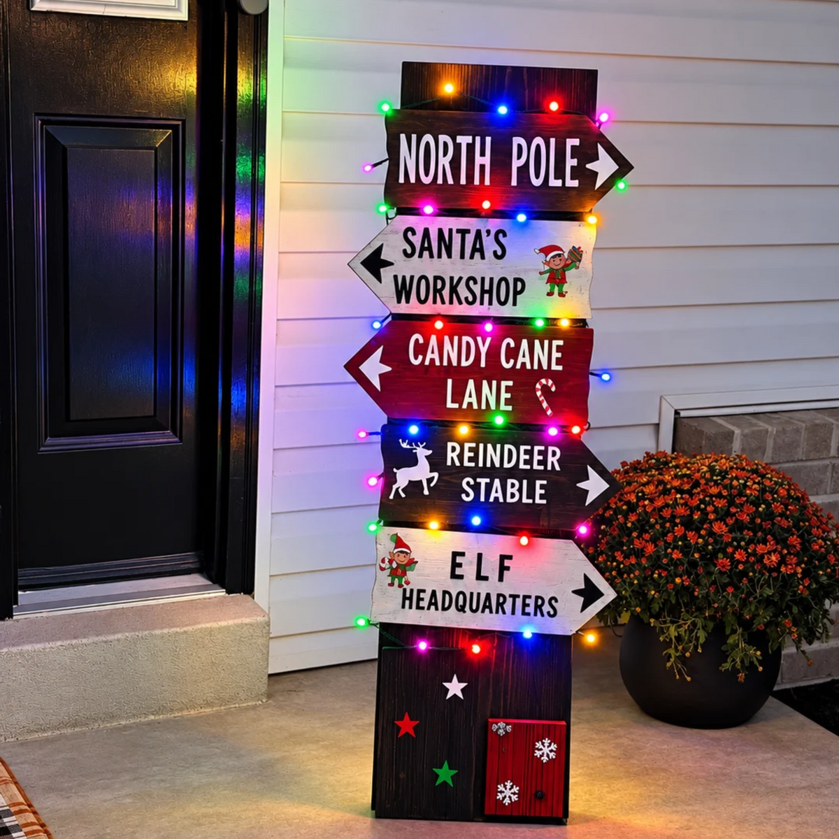 A multi-color lighted vertical wooden Christmas signpost featuring directional signs that read 'NORTH POLE', 'SANTA'S WORKSHOP', 'CANDY CANE LANE', 'REINDEER STABLE', and 'ELF HEADQUARTERS'.