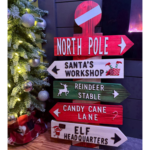 A Christmas-themed wooden signpost with directional arrows pointing to 'NORTH POLE', 'Santa's Workshop', 'Reindeer Stable', 'Candy Cane Lane', and 'Elf Headquarters', styled next to a lit Christmas tree.