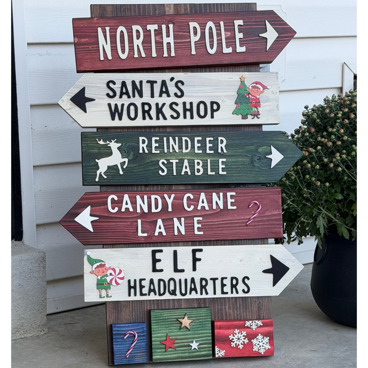 A rustic wooden directional signpost with five arrow-shaped panels reading NORTH POLE, SANTA'S WORKSHOP, REINDEER STABLE, CANDY CANE LANE, and ELF HEADQUARTERS, plus holiday illustrations and wrapped gift accents, photographed outdoors against white siding.
