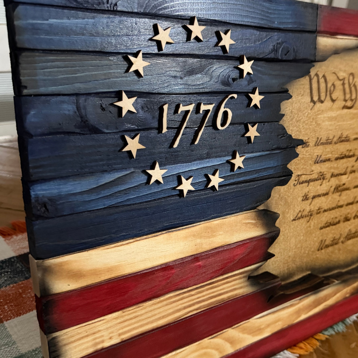 A close-up of a rustic wooden patriotic plaque featuring a distressed American flag design with the year 1776 and a circle of wooden stars, alongside part of a parchment-style United States document.