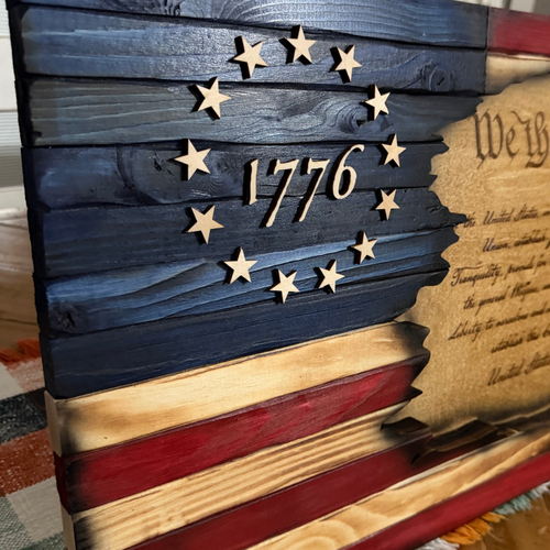 A close-up of a rustic wooden patriotic plaque featuring a distressed American flag design with the year 1776 and a circle of wooden stars, alongside part of a parchment-style United States document.